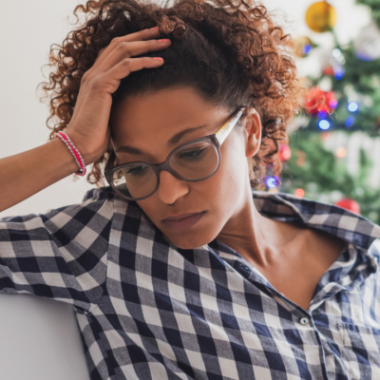 A woman holds her head in one hand. A Christmas tree is behind her. She looks stressed.