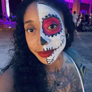 A woman with half of her face painted in Día de los Muertos calavera makeup—white base, black detailing, and a vivid red eye circle—smiles at the camera in a vibrant, festive setting with colorful lights and people celebrating in the background.