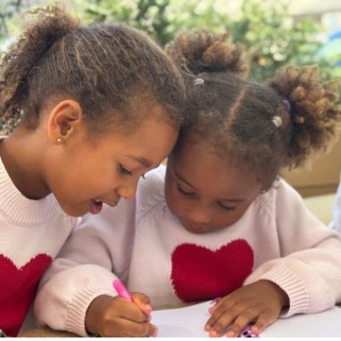 Two young sisters sit close together, writing in a notebook and sharing a quiet, focused moment