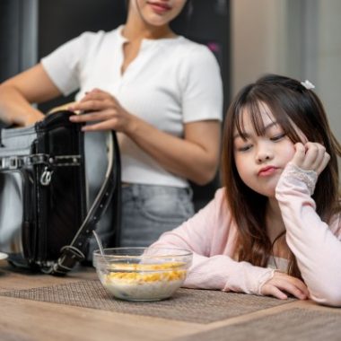 Parent preparing a school bag while anxious child sits at table avoiding breakfast