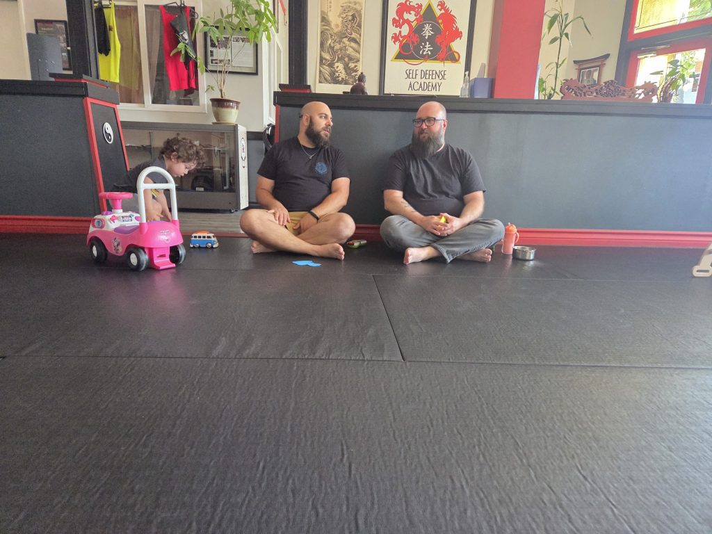 Two dads talking while sitting on a floor with child nearby.