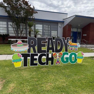 Colorful “Ready, Tech, Go” sign displayed on the lawn outside a school building for a digital literacy event.